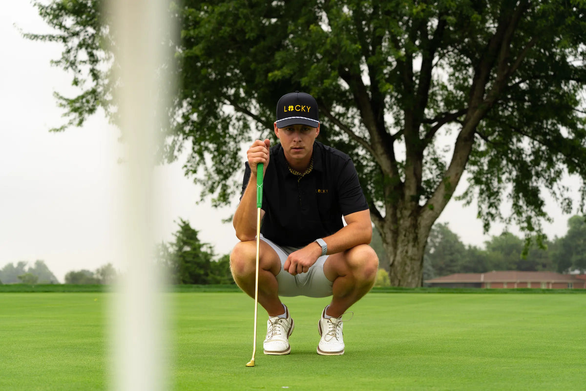 A golfer in a Lucky hat and polo crouched on the green reading a putt, gold putter in hand